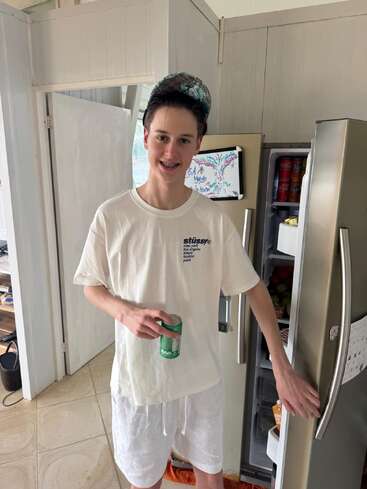 Ein Jugendlicher, der ein weißes Stüssy-T-Shirt, weiße Shorts und eine Mütze trägt, steht lächelnd mit einer Getränkedose in der Hand vor einem offenen Kühlschrank.