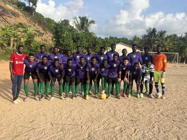 A youth soccer team poses together on a sandy field, wearing matching purple jerseys and green socks, with coaches and goalkeepers standing on each side.