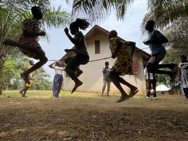 A group of children energetically jump rope outdoors, surrounded by palm trees and a house, enjoying playtime together on a sunny day in a rural setting.
