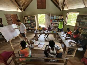 A small classroom scene with a teacher at a whiteboard and students seated at wooden benches, engaged in learning. Bookshelves and natural light brighten the room.