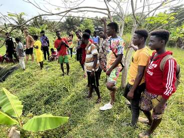 A group of young people gather outdoors on green grass, some standing, some sitting. They appear engaged, possibly listening or participating in an activity under trees.