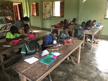 Children sit at wooden desks in a classroom, drawing and coloring. A teacher assists, while bookshelves and windows provide a bright, engaging learning environment.