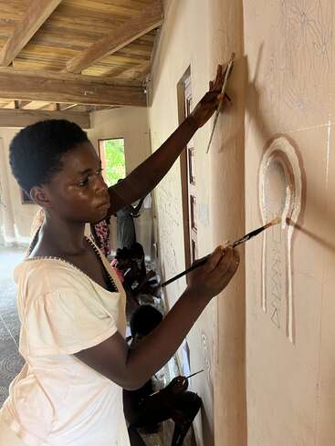 A young woman carefully paints a mural on a light-colored wall in a sunlit room. Several others in the background also work on their own art.