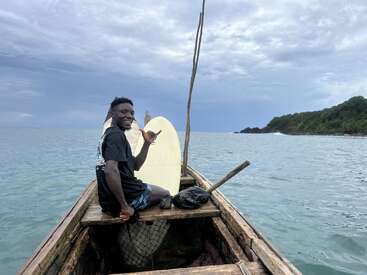 A smiling man sits in a wooden boat with a surfboard, making a shaka sign. The boat is on calm water near a green, hilly shoreline.
