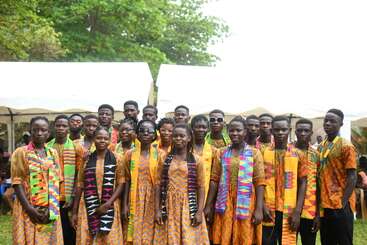 A group of young people in vibrant traditional African attire pose together outdoors, smiling. White tents and lush greenery provide a festive, celebratory backdrop for the gathering.