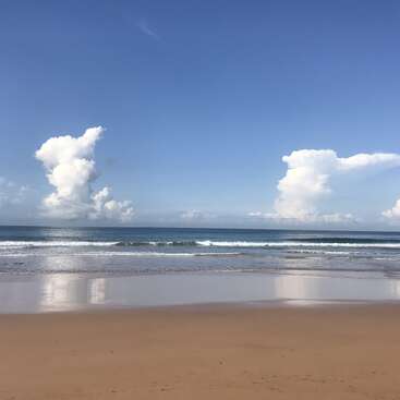 A sandy beach stretches toward a calm ocean under a clear blue sky. Two large fluffy clouds rise in the distance, reflecting gently on wet sand.