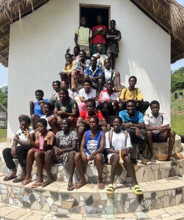 A group of smiling young people sits and stands together on stone steps outside a thatched-roof building, enjoying a sunny day and displaying unity and friendship.