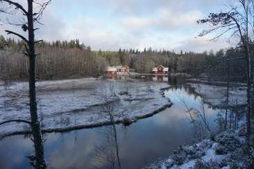Dos casas rojas junto a un río tranquilo y reflectante rodeado de árboles helados y nieve ligera, bajo un cielo azul pálido con nubes dispersas. Una apacible escena invernal.