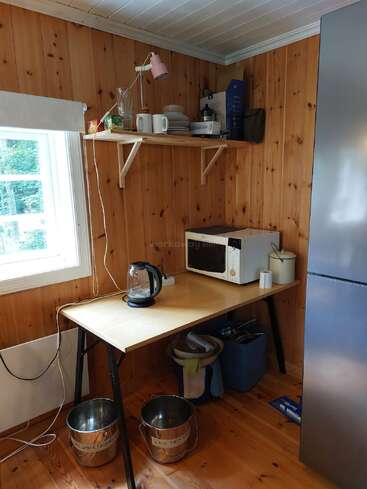 Esta acogedora cocina cuenta con un interior de madera, una mesa con hervidor de agua y microondas, estanterías abiertas con vajilla, dos cubos metálicos etiquetados y una ventana.