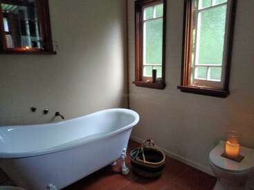 Este acogedor cuarto de baño cuenta con una bañera de patas blancas, dos ventanas esmeriladas, una cesta de mimbre, un inodoro iluminado con velas y una relajante luz natural sobre el suelo de baldosas.