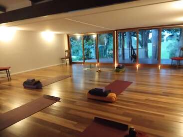 Un sereno estudio de yoga con suelos de madera, esterillas de yoga, bloques y almohadones ordenados. Los grandes ventanales dejan ver la exuberante vegetación del exterior, creando un ambiente tranquilo y acogedor.