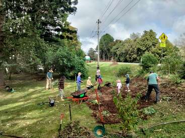 Un grupo de adultos y niños cultivan juntos un huerto cerca de una carretera, utilizando herramientas y carretillas, rodeados de árboles y vegetación bajo un cielo parcialmente nublado.