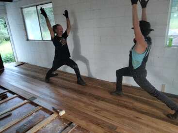 Dos personas adoptan alegremente posturas idénticas a las del yoga sobre un suelo de madera recién instalado en el interior de una luminosa habitación, con guantes y ropa de trabajo, celebrando juntas sus progresos.