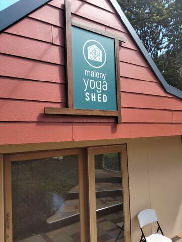 Esta imagen muestra un edificio rojo con un gran cartel verde que dice "Maleny Yoga Shed". Hay una puerta de cristal, una silla blanca plegable y árboles en el exterior.