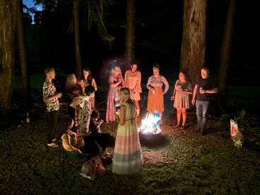 Un grupo de personas se reúne alrededor de una hoguera encendida por la noche en el bosque, hablando y riendo juntos, rodeados de árboles altos y un ambiente relajado.