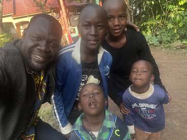 Un hombre y cuatro niños sonríen y posan para un selfie al aire libre. Parecen felices, vestidos con ropa informal, con vegetación y un edificio de fondo.