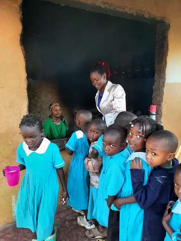 A group of school children in blue uniforms stand in line, waiting for food served by women in a rustic kitchen, one holding a pink cup.