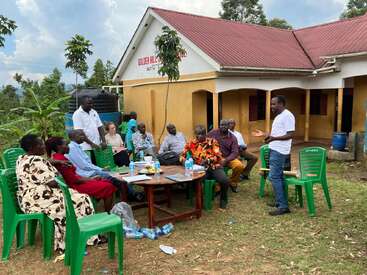 A group of people sit outside, engaged in a discussion led by a standing man. They are gathered near a building called "Golden Hill Academy."