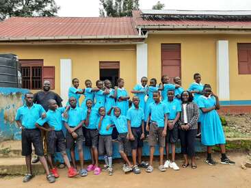 A group of students and two adults pose cheerfully in front of a yellow school building. The students wear matching blue uniforms, displaying camaraderie and happiness.