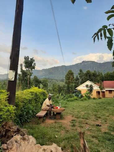An elderly man sits on a wooden bench outdoors, enjoying a meal. Surrounding him are lush greenery, mountains in the background, and a small building nearby.