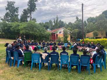 A large group of people sits in a circle on plastic chairs outdoors, engaging in discussion. Green trees, buildings, and cloudy skies form the background scenery.