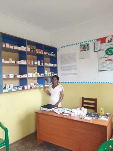 A woman stands in a small pharmacy or clinic, surrounded by shelves of medication, a wooden desk with files, medicine boxes, and medical supplies, bright atmosphere.
