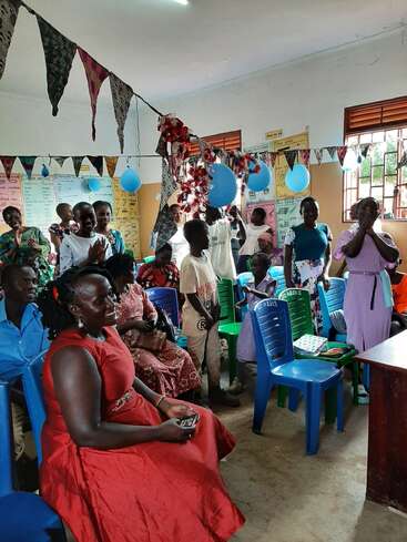 A group of people, including children and adults, gather in a decorated room with balloons and bunting, celebrating together with smiles, clapping, and joyful expressions.