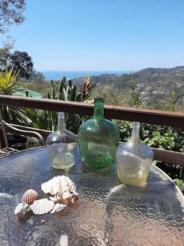 Das Bild zeigt eine ruhige Umgebung im Freien mit einem Glastisch, Muscheln und Glasflaschen, mit Blick auf eine malerische Landschaft mit Hügeln, Pflanzen und einem entfernten Gewässer unter einem klaren blauen Himmel.