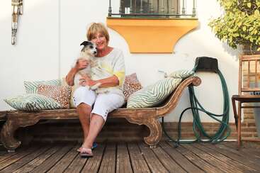 Das Bild zeigt eine Frau mit langen Haaren und einem weißen Hemd, die mit gekreuzten Beinen auf dem Boden sitzt, umgeben von einer natürlichen Umgebung mit Bäumen und einem Gewässer.