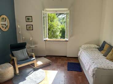Habitación luminosa y acogedora con una ventana abierta, cama individual, sillón moderno, puf, alfombra redonda, cojines, mesita auxiliar, cuadro en la pared y una exuberante vegetación en el exterior.