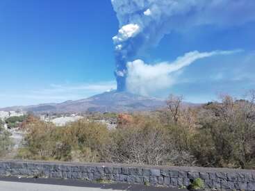 Um vulcão está em erupção, lançando uma enorme coluna de cinzas e fumaça no céu azul brilhante. Dá pra ver arbustos, árvores e um muro de pedra.