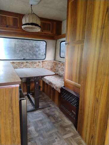 Esta imagem mostra o interior aconchegante de uma caravana vintage. Ela tem painéis de madeira, uma mesinha, almofadas estampadas, armários suspensos e janelas grandes.