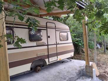 A imagem mostra uma caravana antiga em tons de bege e marrom estacionada sob um abrigo de madeira em uma área arborizada, cercada por árvores verdes e galhos frondosos.