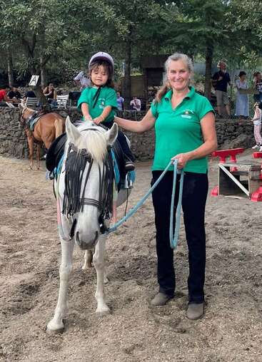 Uma mulher sorridente segura as rédeas de um cavalo branco, enquanto uma criança pequena, usando um capacete, está montada no cavalo. Os dois vestem camisetas verdes iguais. Cenário ao ar livre.