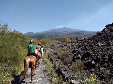 Das Bild zeigt drei Menschen, die auf Pferden auf einem Steinweg reiten, umgeben von üppigem Grün und schroffen vulkanischen Felsformationen vor einer majestätischen Bergkulisse.