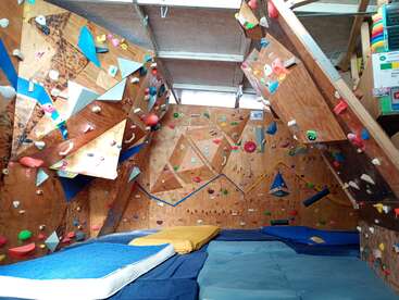 This image shows an indoor bouldering gym with wooden climbing walls, colorful holds, geometric volumes, and padded mats on the floor for safety and comfort.