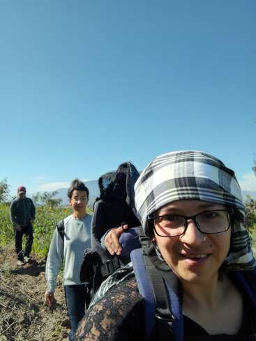 Three people are hiking outdoors under a clear blue sky. The woman in front wears glasses and a checkered headscarf, smiling. The group looks cheerful and adventurous.