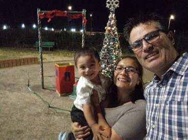 A happy family of three smiles together at night, posing near a decorated Christmas tree and festive playground, celebrating the holiday season outdoors with joy.