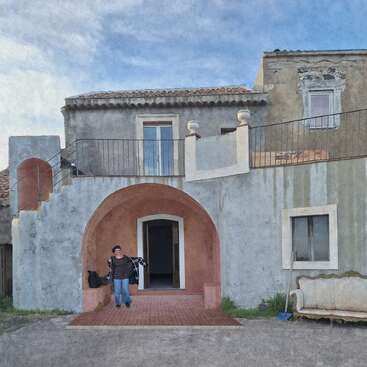 A person stands under a pink arch of an old, rustic two-story building with weathered walls, outdoor staircase, balcony, and an antique sofa outside. Blue sky.
