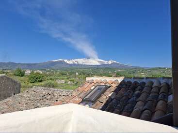 Terracotta rooftops in the foreground, green countryside beyond. Snow-capped volcano with smoke plume rises under a clear blue sky, creating a stunning, peaceful landscape scene.