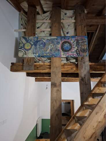 A rustic wooden staircase leads to a loft area with exposed beams. An abstract, colorful painting hangs above, against a backdrop of patterned fabric. Cozy interior.