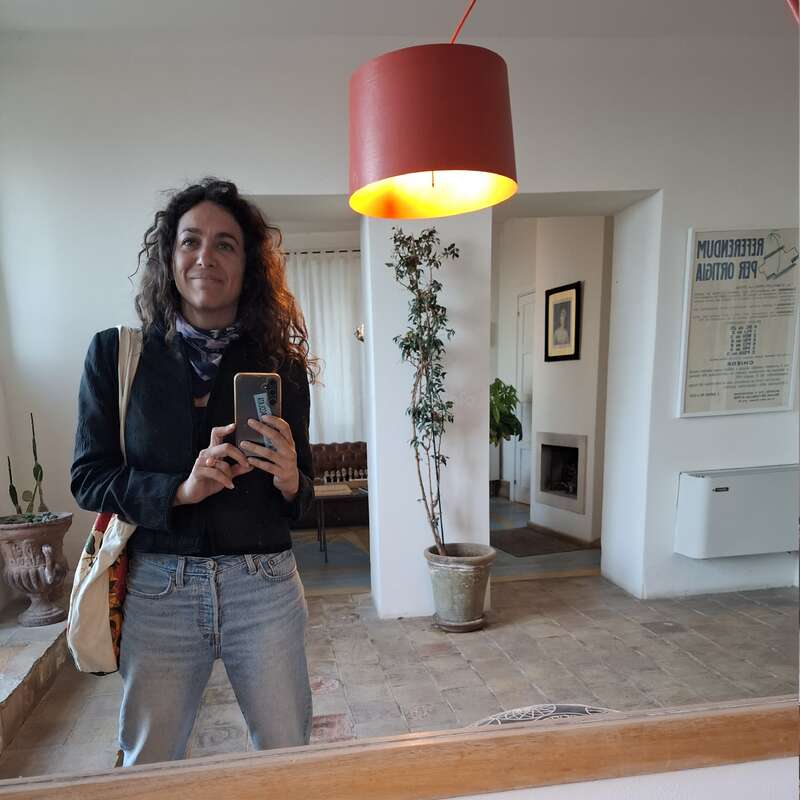 A woman with curly hair takes a mirror selfie in a cozy, stylish room. She smiles, wearing jeans and a scarf, with plants and art in the background.