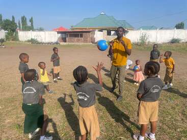 Ein Mann in einem gelben Hemd spielt mit einem blauen Ball, umgeben von Kindern auf einer Wiese, mit einem Gebäude und einem Zaun im Hintergrund an einem sonnigen Tag.