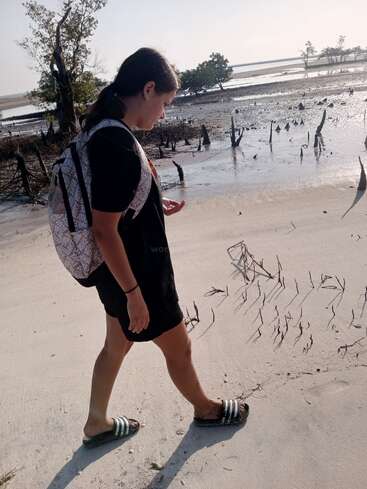 Eine Frau geht mit einem Rucksack, schwarzen Shorts und Sandalen an einem Sandstrand spazieren, mit einem Mangrovenwald im Hintergrund und einem Gewässer in der Ferne.