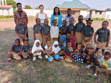 Das Bild zeigt eine Gruppe von Kindern und Erwachsenen, die auf einer Wiese für ein Foto posieren, während im Hintergrund ein Haus und Gebäude zu sehen sind.