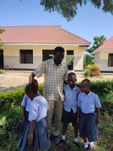 Das Bild zeigt einen Mann, der mit vier Kindern vor einem Gebäude steht, umgeben von üppigem Grün und einem klaren blauen Himmel.