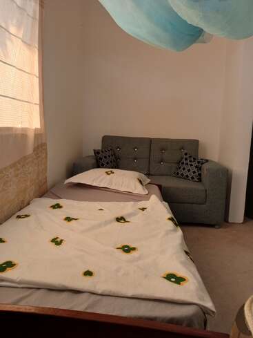 Das Bild zeigt ein kleines Zimmer mit einem Bett, einer Couch und einem Fenster, auf dem eine weiße Bettdecke mit grünen und gelben Mustern und eine graue Couch mit zwei Kissen zu sehen sind.