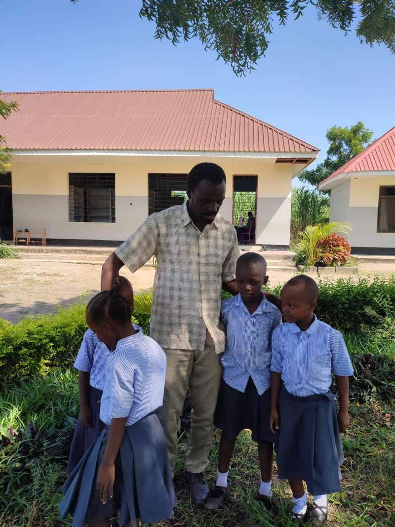 Das Bild zeigt einen Mann, der mit vier Kindern vor einem Gebäude steht, umgeben von üppigem Grün und einem klaren blauen Himmel.