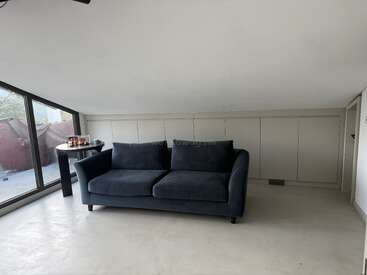 Une pièce moderne et minimaliste avec un plafond incliné, comprenant un canapé gris foncé centré contre le mur et une petite table d'appoint près de grandes fenêtres.