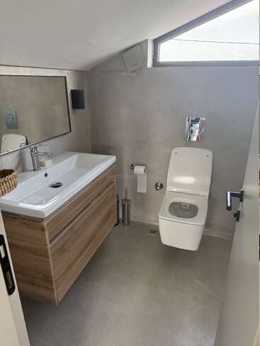 Cette salle de bains moderne se compose de toilettes murales, d'un meuble-lavabo en bois épuré avec évier, d'un grand miroir, d'un plafond incliné, de la lumière naturelle d'une fenêtre triangulaire et d'un décor minimaliste.